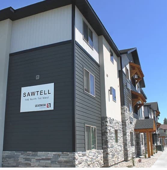 Modern three-story apartment building in Rexburg with stone and dark gray siding, white upper walls, and wood accents. A sign reads “SAWTELL 146 North 1st West.” The sky above Sawtell Rexburg is clear and blue.