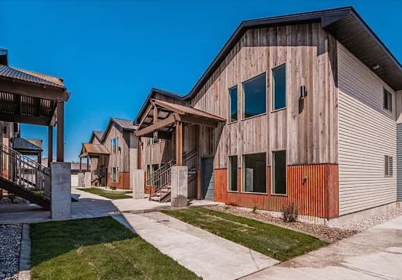 Modern wooden apartment buildings with large windows, covered entryways, and metal siding accents define Teton River Lofts Rexburg. A concrete walkway and green lawns run alongside the buildings under a clear blue sky.
