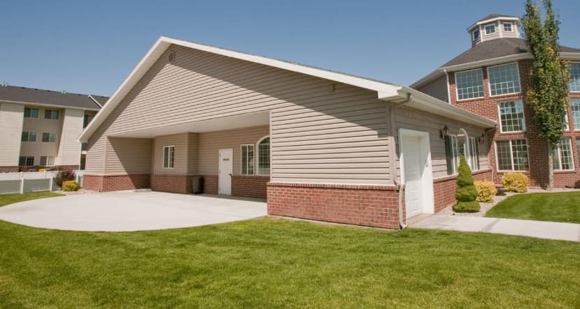 A single-story beige building with red brick accents and a covered entryway sits on a neatly manicured lawn at The Village Rexburg, with a multi-story brick building visible in the background under a clear blue sky.