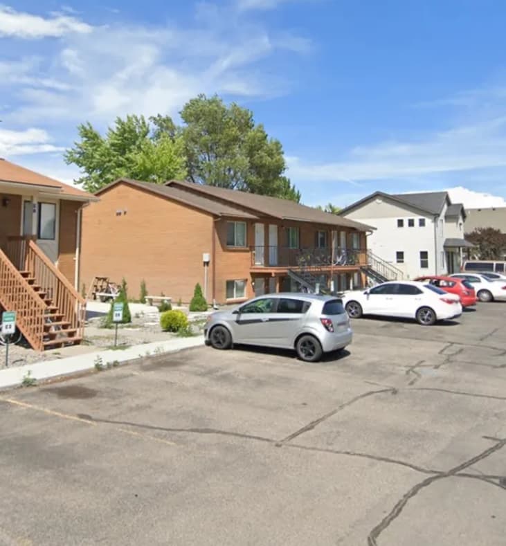 A small apartment building, Wayside Manor Rexburg, features a brown brick exterior and wooden stairs beside a parking lot with several cars on a sunny day. Trees and a white building can be seen in the background.