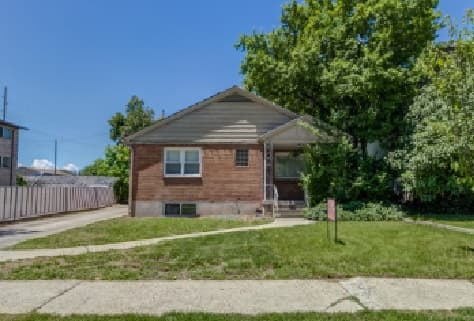 A small, single-story brick house with a gabled roof, a driveway on the left, and a well-kept lawn. Surrounded by trees and shrubs under a clear blue sky, it evokes the cozy charm of a Yearsley Duplex Provo home.