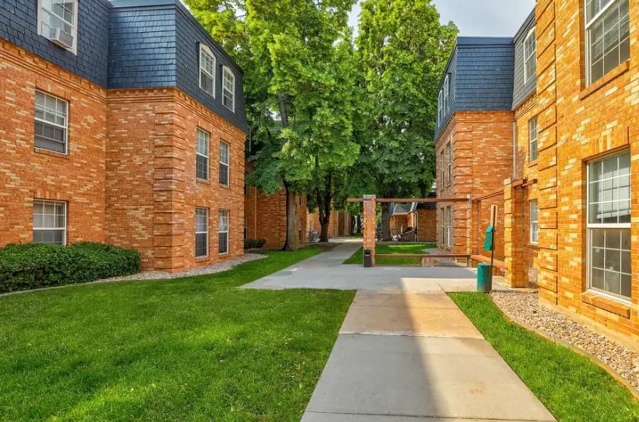 A walkway runs between two brick apartment buildings at Eleven13 Apartments Fort Collins, with manicured lawns, trees, and a covered seating area in the background on a sunny day.