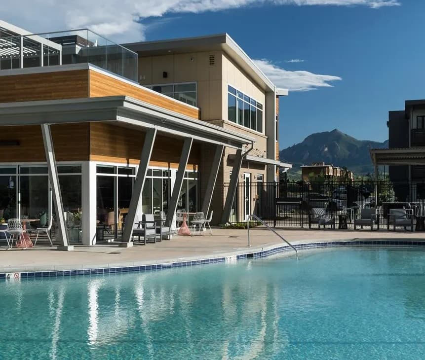 Mysa at the Crossing Boulder features a modern building with large windows and wooden accents, overlooking a clear outdoor swimming pool surrounded by chairs and tables, with mountain views and a blue sky dotted with clouds in the background.