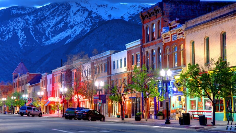 Colorful storefronts and lit street lamps line a lively small-town street at dusk in Ogden, with parked cars and trees along the sidewalk. Snow-capped mountains rise dramatically behind Aubrey Apartments in Ogden.