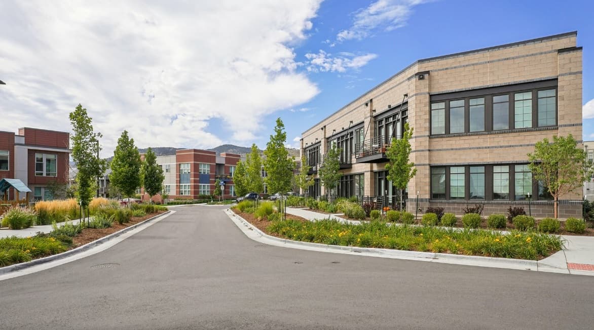 Modern office buildings with large windows line a clean, paved street near The Armory Apartments Boulder, surrounded by landscaped greenery, small trees, and flower beds under a partly cloudy sky.