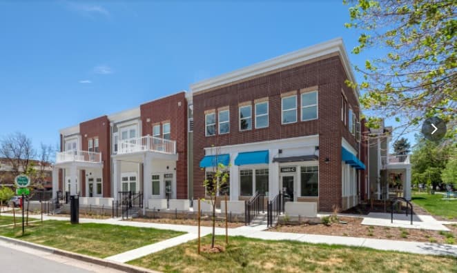 Attention Home Apartments Boulder: A modern, two-story brick building with large windows and blue awnings, featuring balconies and small trees along the sidewalk, under a clear blue sky.