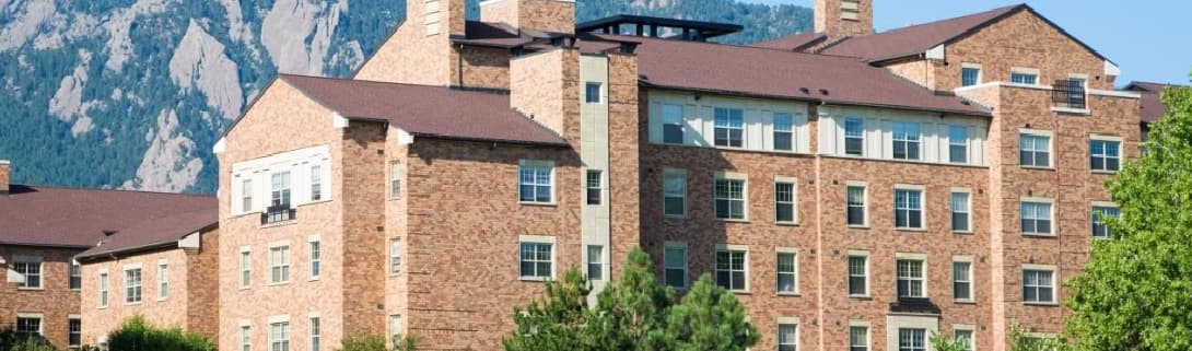 A large brick building with many windows, Bear Creek Apartments B Boulder sits against a backdrop of green trees and rocky mountains under a clear blue sky.