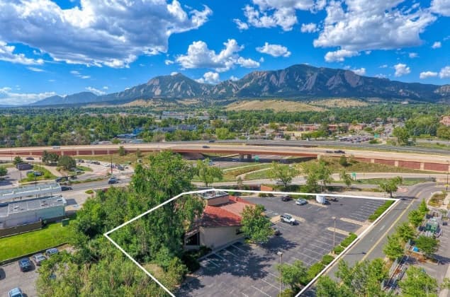 Aerial view of Boulder Housing Boulder building and parking lot outlined in white, with a highway, green trees, and mountains in the background under a blue sky with scattered clouds.