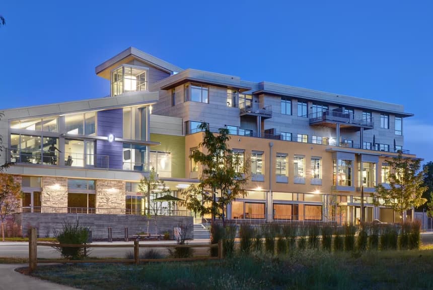 A modern three-story apartment building, Block One Apartments Fort Collins, features large windows and balconies illuminated at dusk. The exterior boasts a mix of materials and colors, with landscaping and a wooden fence in the foreground.
