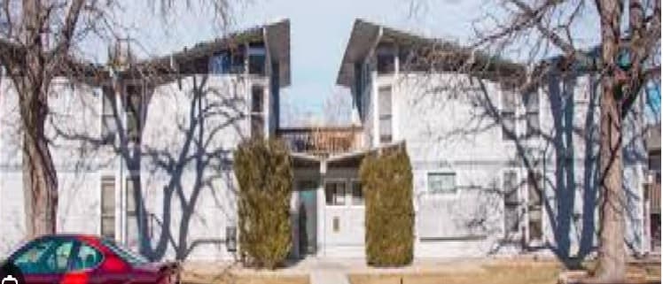 Blue Spruce Apartments Boulder: A modern white duplex building with slanted roofs, tall windows, and two bushy trees at the entrance. A red car is parked on the street in front, framed by leafless trees.