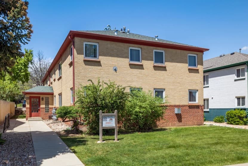 Two-story beige brick Boulder Apartments building with red trim, surrounded by green grass, shrubs, and a sidewalk. A white sign with red lettering stands near the entrance under a clear blue sky in Boulder.