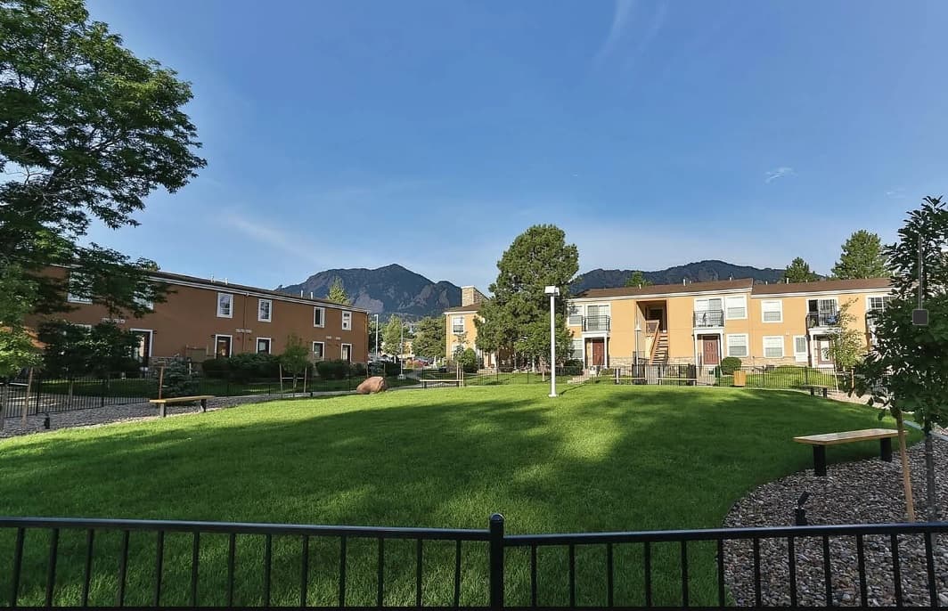 A green lawn with benches is surrounded by brown apartment buildings, trees, and a black metal fence at Boulder Creek Apartments Boulder, with mountains and a clear blue sky in the background.