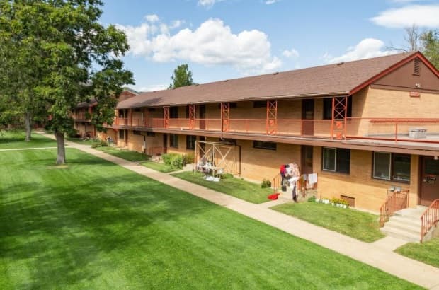 A two-story brick Boulder Trio Boulder apartment building with orange railings faces a large, well-maintained green lawn and a sidewalk on a sunny day. Some balconies and ground areas display personal items.