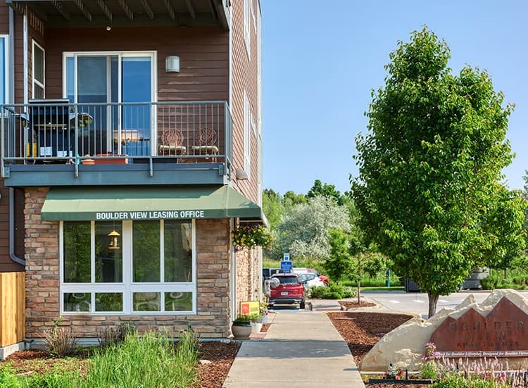 Two-story building with a green awning reading Boulder View Leasing Office above the entrance; balcony and potted plants above; sidewalk, tree, and Boulder View Apartments Boulder sign to the right.