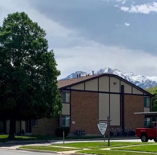 Bramblewood Apartments Ogden features a two-story brick building with brown trim, set before a large green tree. Snow-capped mountains and a partly cloudy sky rise in the background, with sidewalk and grass in the foreground.
