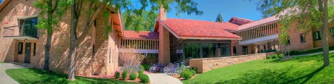 A sunny day at Buckingham Hall Boulder, a brick building with a red-tiled roof, large windows, and a courtyard filled with trees, green grass, and bicycles parked by the entrance.