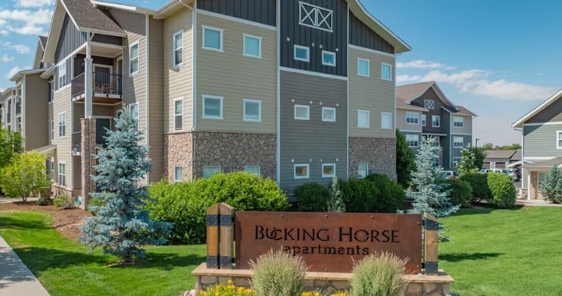 A modern apartment complex with tan and gray siding, stone accents, green lawns, shrubs, and trees. In front is a sign reading Bucking Horse Apartments Fort Collins. Bright, sunny weather.