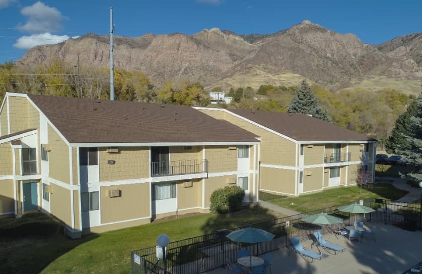 Two beige apartment buildings with brown roofs stand before rocky mountains at Canyon Crest Apartments Ogden. In the foreground, a fenced patio area with tables, chairs, and umbrellas sits among trees with autumn leaves under a blue sky.
