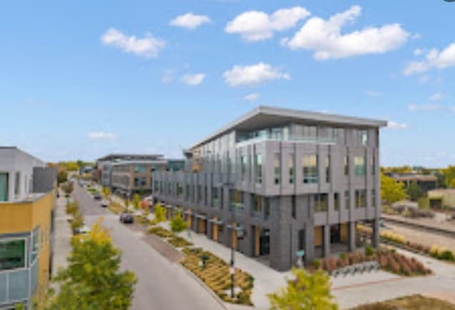 A modern, multi-story office building at Boulder Commons features large windows and landscaped greenery, with cars parked nearby and other commercial offices and retail in Boulder under a partly cloudy sky.