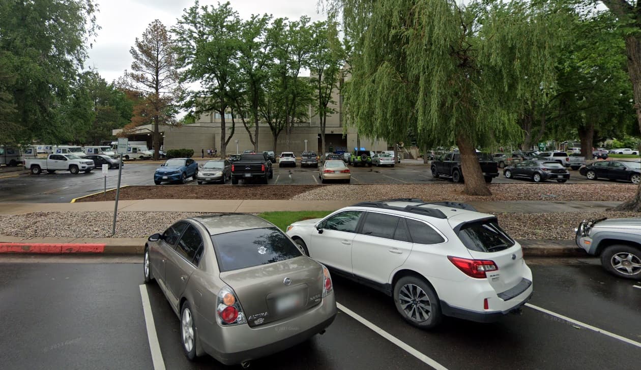 A parking lot with several parked cars, trees with green leaves, and the large CSU LOFTS Fort Collins building in the background under an overcast sky. The ground appears wet, likely from recent rain.