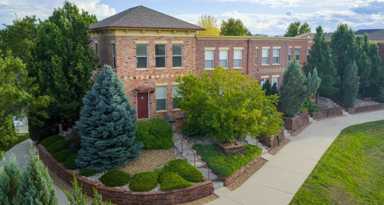 A two-story brick building with many windows at Enclave Rigden Farm Townhomes, Fort Collins, surrounded by lush trees and gardens. A red door and steps lead to the entrance, with stone borders accenting the well-maintained landscaping.