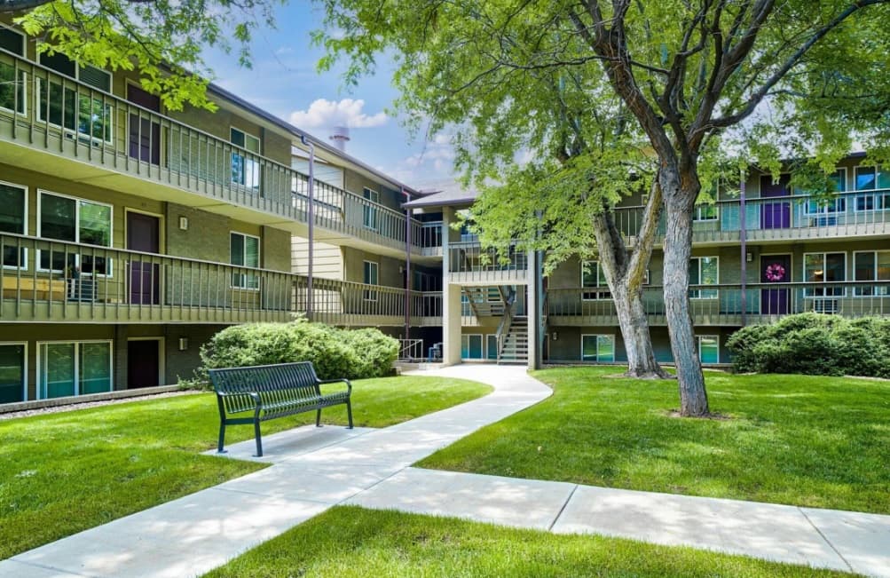 The courtyard at Flatiron View Apartments Boulder features green grass, a sidewalk, metal bench, lush bushes, and mature trees. Surrounding buildings with balconies offer multiple floors. The partly cloudy sky adds to the serene atmosphere.