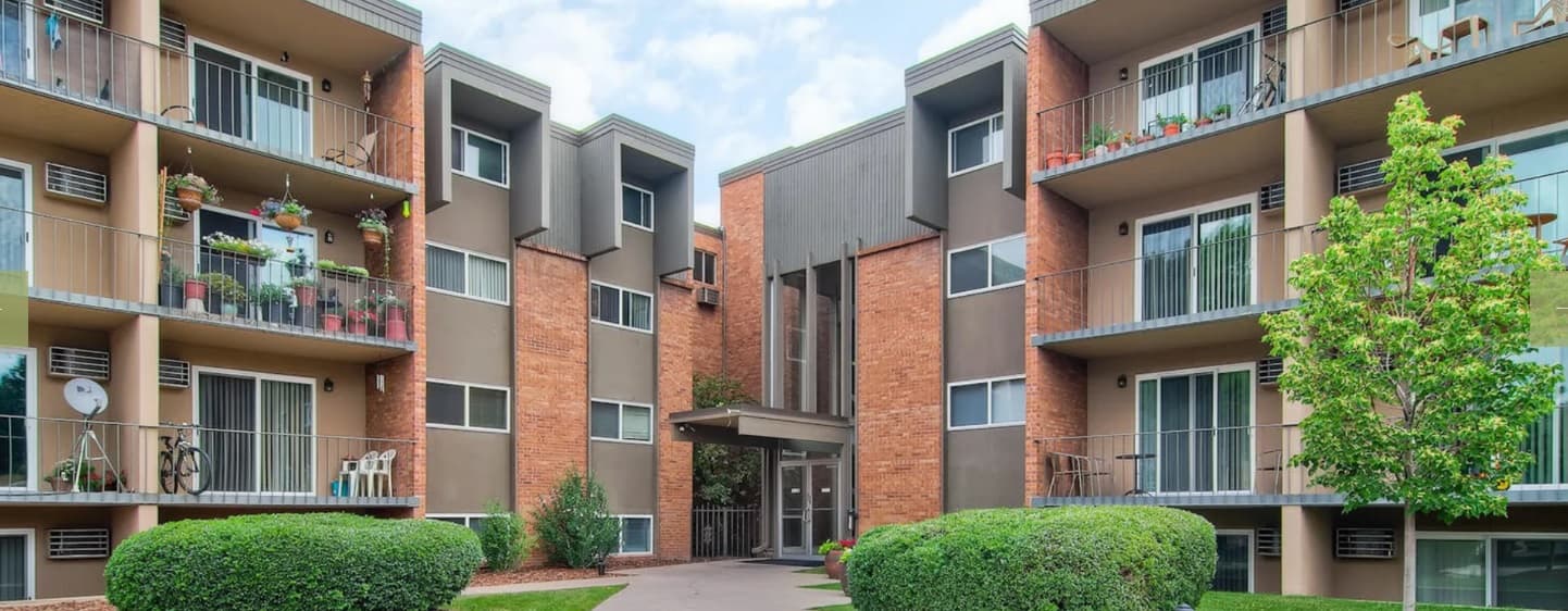 GlenLake Apartments Boulder features a modern three-story building with a brick and gray exterior, balconies adorned with plants and furniture, and well-kept bushes and trees out front.