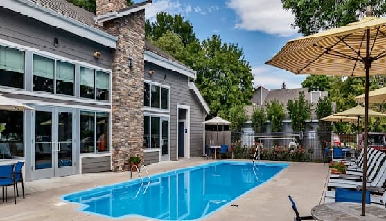 Outdoor swimming pool beside a modern gray building with large windows at Governor's Park Fort Collins, surrounded by lounge chairs, yellow umbrellas, and trees under a partly cloudy sky.