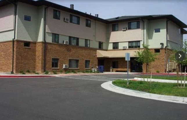 Harmony Road Apartments Fort Collins is a three-story building with brick on the lower level and light siding on the upper floors, featuring several windows, an entrance canopy, and a paved driveway in front.