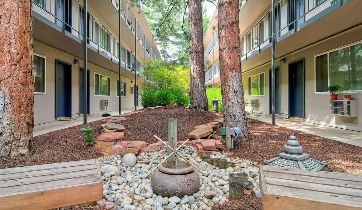 Nestled between Harper House Apartments Boulder, this tranquil courtyard features tall trees, benches, rocks, and a small Japanese-style stone fountain, creating a peaceful garden setting.