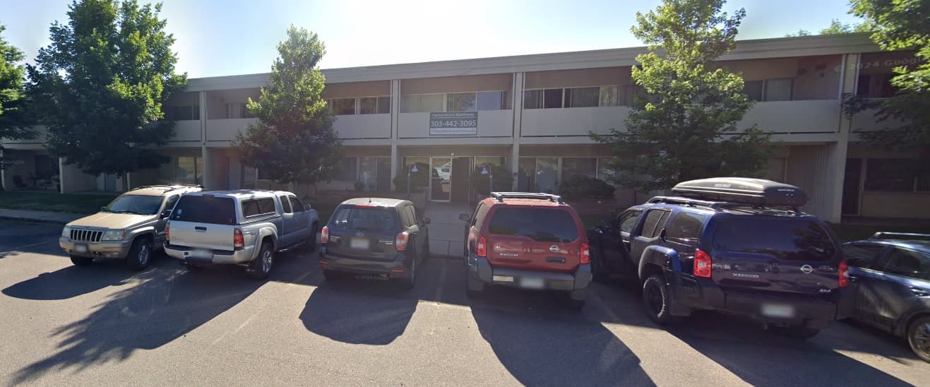 A two-story office building with large windows, neighboring Harvest Manor Apartments Inc Boulder, and a row of five parked SUVs and cars in front, partially shaded by leafy trees on a sunny day.
