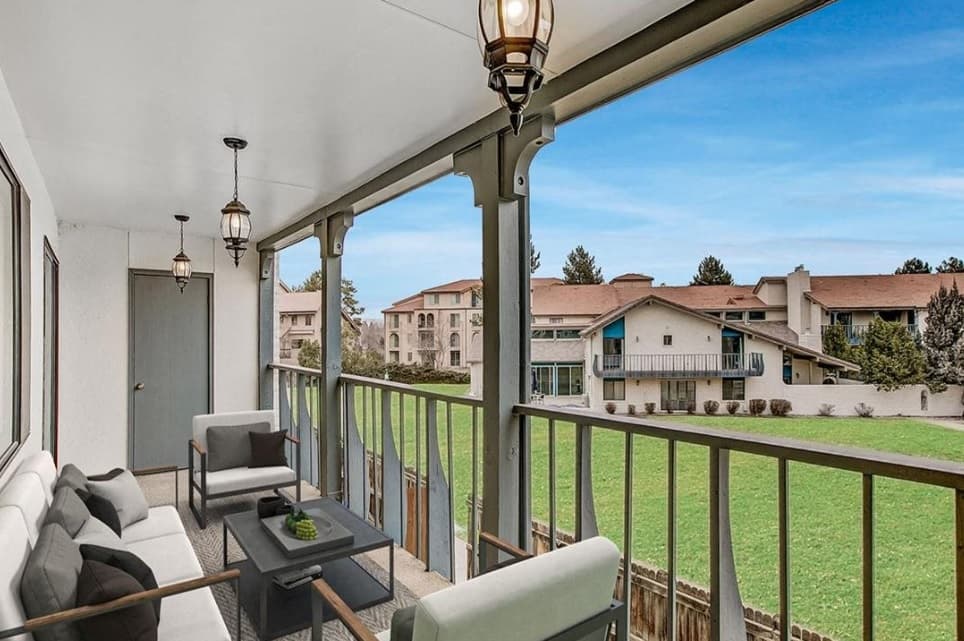 A covered balcony with cushioned seating and a coffee table at Henley and Remy Apartments Boulder overlooks a grassy area and neighboring residential buildings under a clear blue sky.