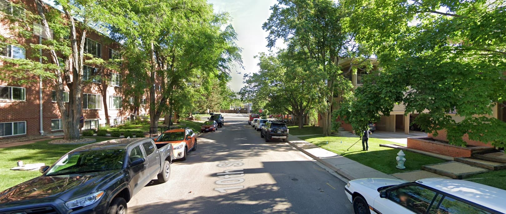 A leafy, residential street lined with parked cars and apartment buildings like Imperial House Apartments Boulder. Tall green trees provide shade, and a few people walk on the sidewalks under sunny skies.