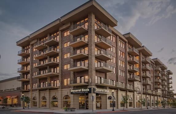 A multi-story brick apartment building with balconies and large windows, Junction View Apartments Ogden features a Deseret Book store on the ground floor at the corner of a city street in the evening.