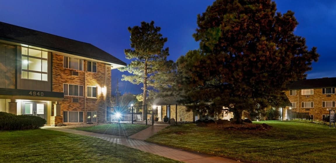 A well-lit apartment building exterior at dusk, with large windows, brick walls, tall trees, and neatly trimmed grass in the foreground. A sidewalk leads toward the Kimberly Court Apartments Boulder entrance.