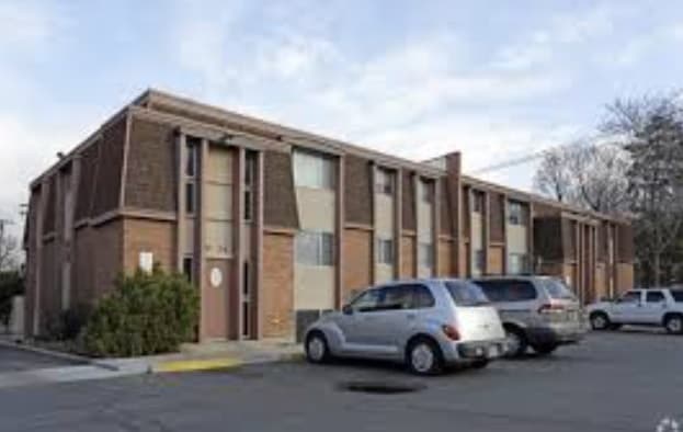 A two-story brick apartment building with brown trim, several windows, and parked cars in front of it at Kingstowne Apartments Ogden, all beneath a clear sky.