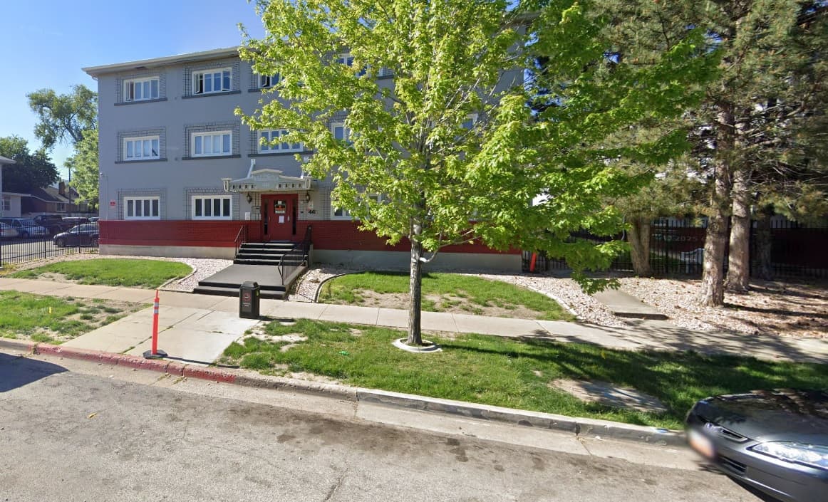 A three-story gray and red apartment building, managed by La Porte Management Ogden, with a tree in front, steps to the main entrance, and a sidewalk along a quiet street on a sunny day.
