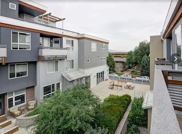 Modern apartment complex, Landmark Lofts Boulder features gray and white exteriors, multiple balconies, large windows, and a shared outdoor patio area with seating—surrounded by green trees and parked cars in the background.