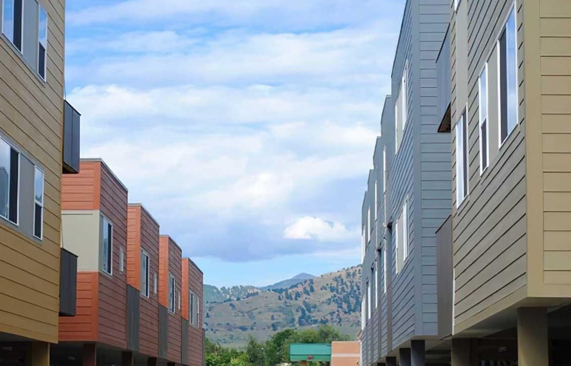 A view between two rows of modern apartment buildings at Ledges Apartments Boulder, with a distant mountain and a blue sky with clouds in the background.