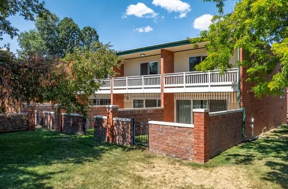 Madeline Creek Apartments Boulder features a two-story brick building with white railings, balconies, and a fenced courtyard, all set amid green grass, trees, and a blue sky with clouds.