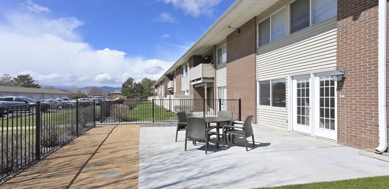 A fenced patio with a table and four chairs sits outside Meadow Creek Apartments Boulder, a two-story building with brick and beige siding. Under a partly cloudy sky, grass and a walkway border the inviting outdoor space.
