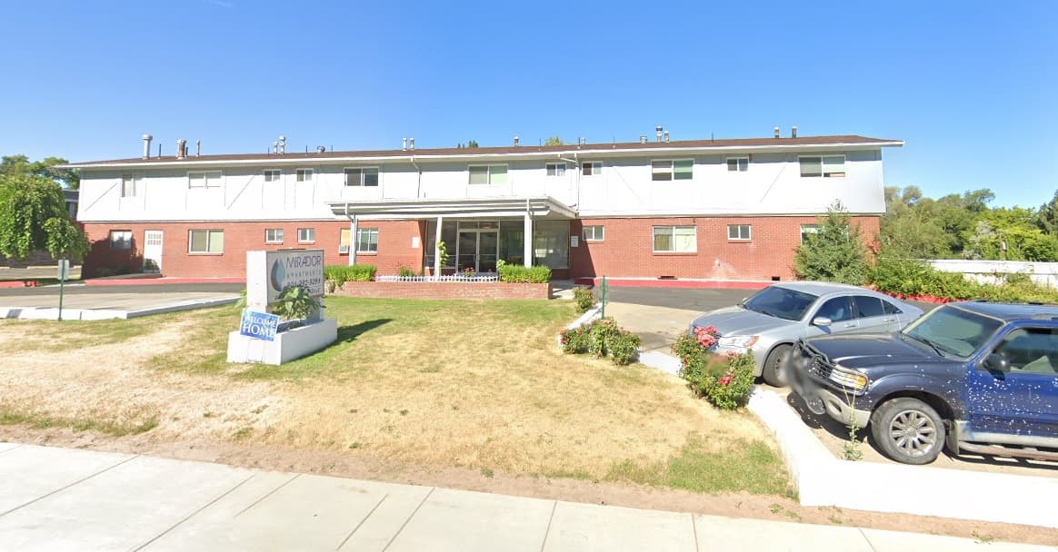 A two-story apartment building with red brick on the lower level and white siding above. Two cars are parked in front, and a sign labeled Moody stands on a small patch of grass near the Mirador Apartments Ogden sidewalk.