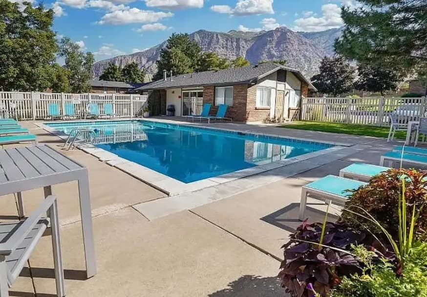 A rectangular outdoor pool at Mountain Ridge Manor Ogden is surrounded by lounge chairs and tables, with a small building and trees nearby. Mountains and a partly cloudy sky are visible in the background.