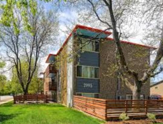 Nest On 30th Boulder is a modern three-story apartment building with a brick and blue exterior, large windows, red roof accents, and wooden fencing, surrounded by trees and green grass on a clear day.