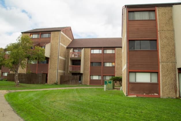 The Newton Court Building 2J in Boulder is a three-story apartment building with brown and beige paneling, large windows, and a sloped roof, surrounded by a grassy lawn and a few trees under a cloudy sky.