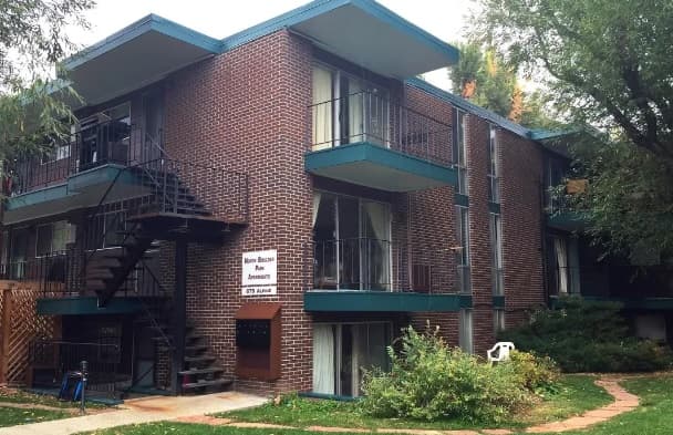 A two-story brick building, North Boulder Park Apartments Boulder, features teal trim, outdoor staircases, balconies, glass doors, and a sign near the entrance. Surrounded by green grass, trees, and a winding path.