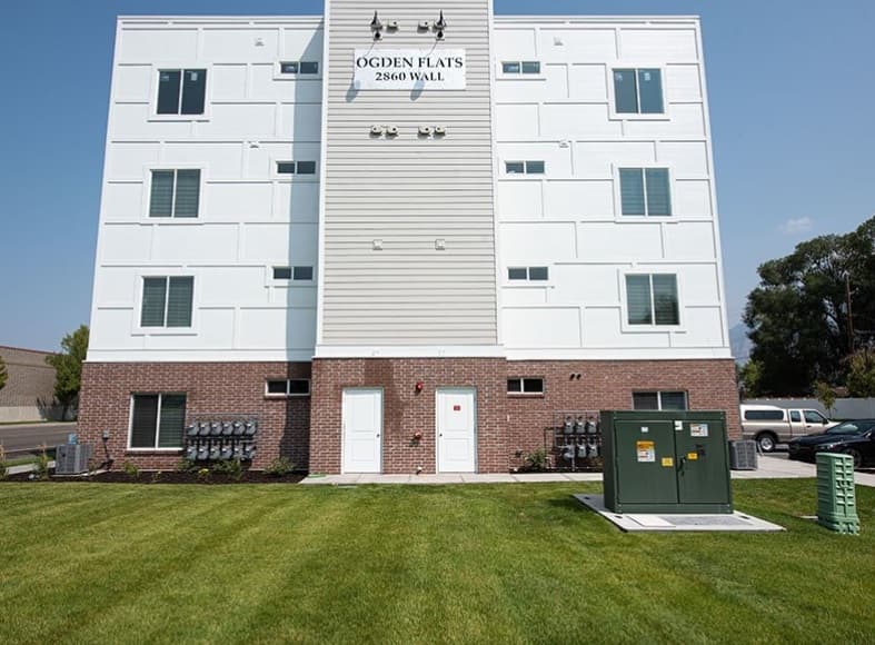 Ogden Flats in Ogden is a modern four-story apartment building at 2860 Wall, featuring a sleek brick and white exterior and a neatly mowed green lawn. Electrical boxes and utility equipment are visible in the foreground.