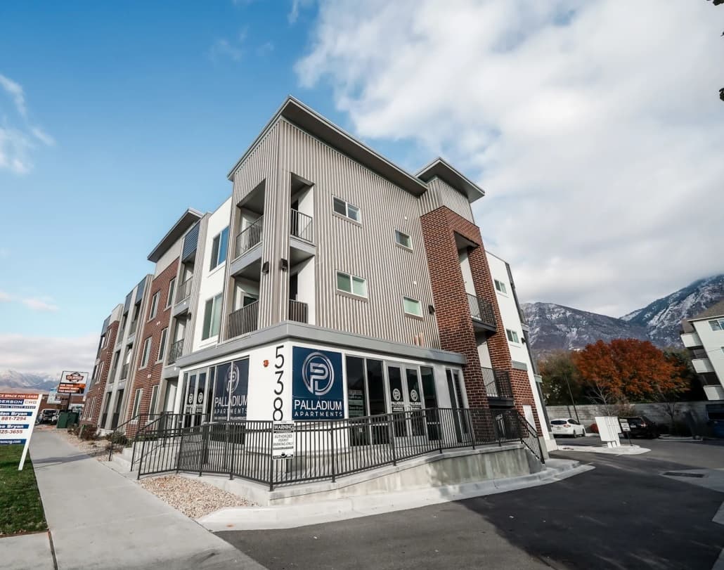 A modern three-story apartment building with brick and light siding, large windows, balconies, and a sign reading Palladium Apartments Provo 5738. Sidewalk, parking lot, and mountains visible in the background.