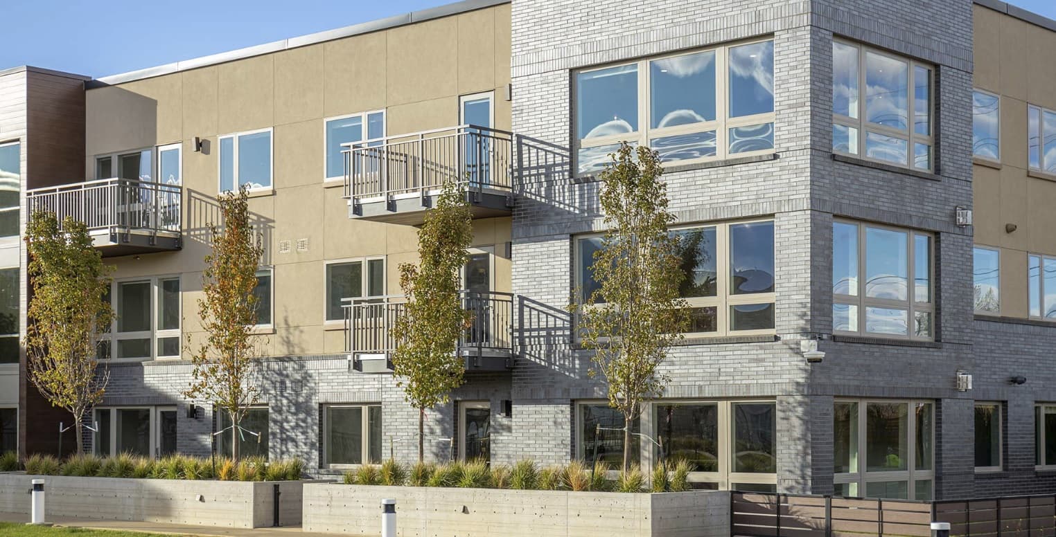 A modern three-story apartment building, reminiscent of Parc Mosaic Apartment Homes Boulder, with large windows, small balconies, and young trees in front. The exterior blends gray brick and tan panels under a clear blue sky.