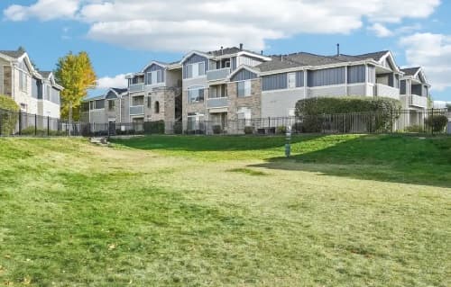 The Pavilions at Silver Sage Fort Collins is a large apartment complex with beige and gray siding, multiple balconies, and large windows, surrounded by a well-maintained grassy lawn and some trees under a partly cloudy blue sky.